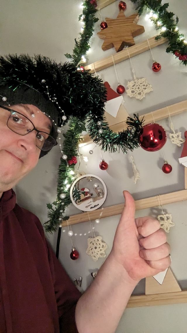 Ein albernes Selfie von mir. Ich habe einen künstlichen Weihnachtsbaum auf dem Kopf, der mir schlapp vom Kopf herunterhängt mit einer roten Kugel daran. Ich presse die Lippen aufeinander, halte den Daumen sarkastisch nach oben, im Hintergrund ein weihnachtlich geschmücktes Wandgehänge in Dreieckform mit verschiedenem weihnachtlichen Zeug. Der Daumen nach oben ist bezogen auf Weihnachten, auf das Fest und die Vorbereitungen.